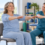 Mujer realizando movimientos suaves de brazos junto a una kinesióloga en un centro de rehabilitación.